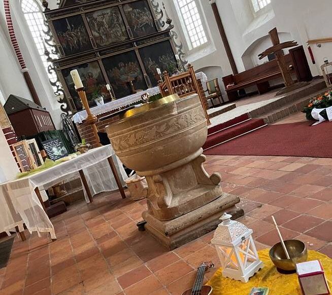 Blick in die Kirche Victorbur, zu sehen ist der Altar, das Taufbecken und davor liegen auf einer kleinen Decke auf dem Boden eine kleine Gitarre, eine Kerzenlaterne, eine Klangschale und ein beschriftetes und bemaltes Kreuz.